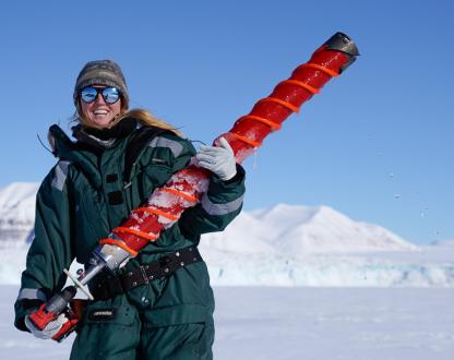 Female polar marine biologist stands in a snowy landscape holding an oversized drill bit