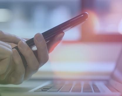 Photo of a person on their laptop holding a mobile phone. 