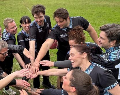 A group of DISR staff belonging to the DISR touch football team stand in a circle on an oval. They are wearing black shirts with a blue artwork celebrating NAIDOC Week. The players are standing in circle with their hands joining in the centre for a hand stack.