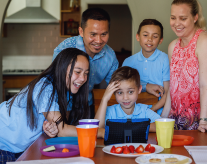 Two parents and their 3 children are smiling while they stand and sit around their dining table as they talk to family overseas using a tablet.