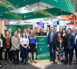 A group of people in business attire standing at a large, open plan exhibition stand at the 2023 World Mining Congress. The stand displays the Australian Government’s green and gold ‘Nation Brand’, including a kangaroo and decorative elements representing Australia’s rivers and bush tracks.