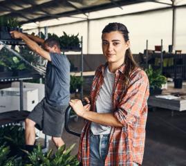 A woman in a small warehouse leans against some equipment. A man behind her lifts a tray of plants off a shelf.