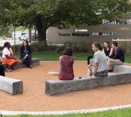 Eight female staff and a man are sitting on wooden sleepers as part of a yarning circle. In the  background, a wall has Geoscience Australia written on it, and the words 'Yuuma Welcome'.