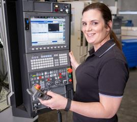 Image shows a woman in a factory. She is standing next to the control panel for a machine tool, smiling and looking towards the camera.